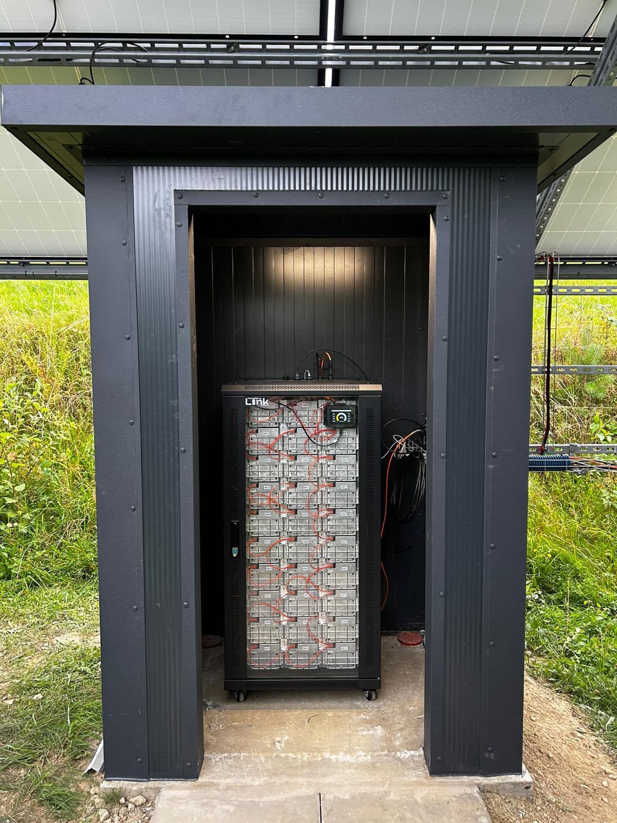 Szafa rack z modułami bateryjnymi w obudowie zewnętrznej, ustawiona pod naziemną instalacją fotowoltaiczną